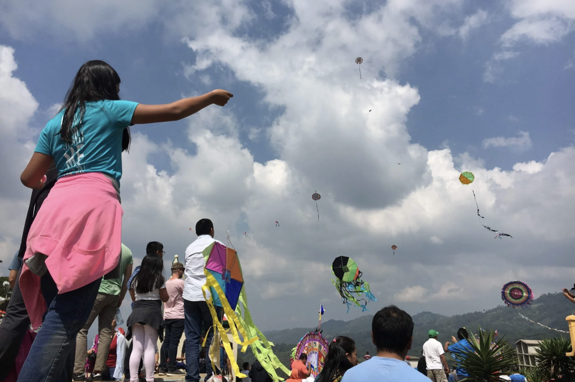 A girl is holding the string to a kite that is flying in the sky with others at the Kite Festival in Sumpango, Guatemala