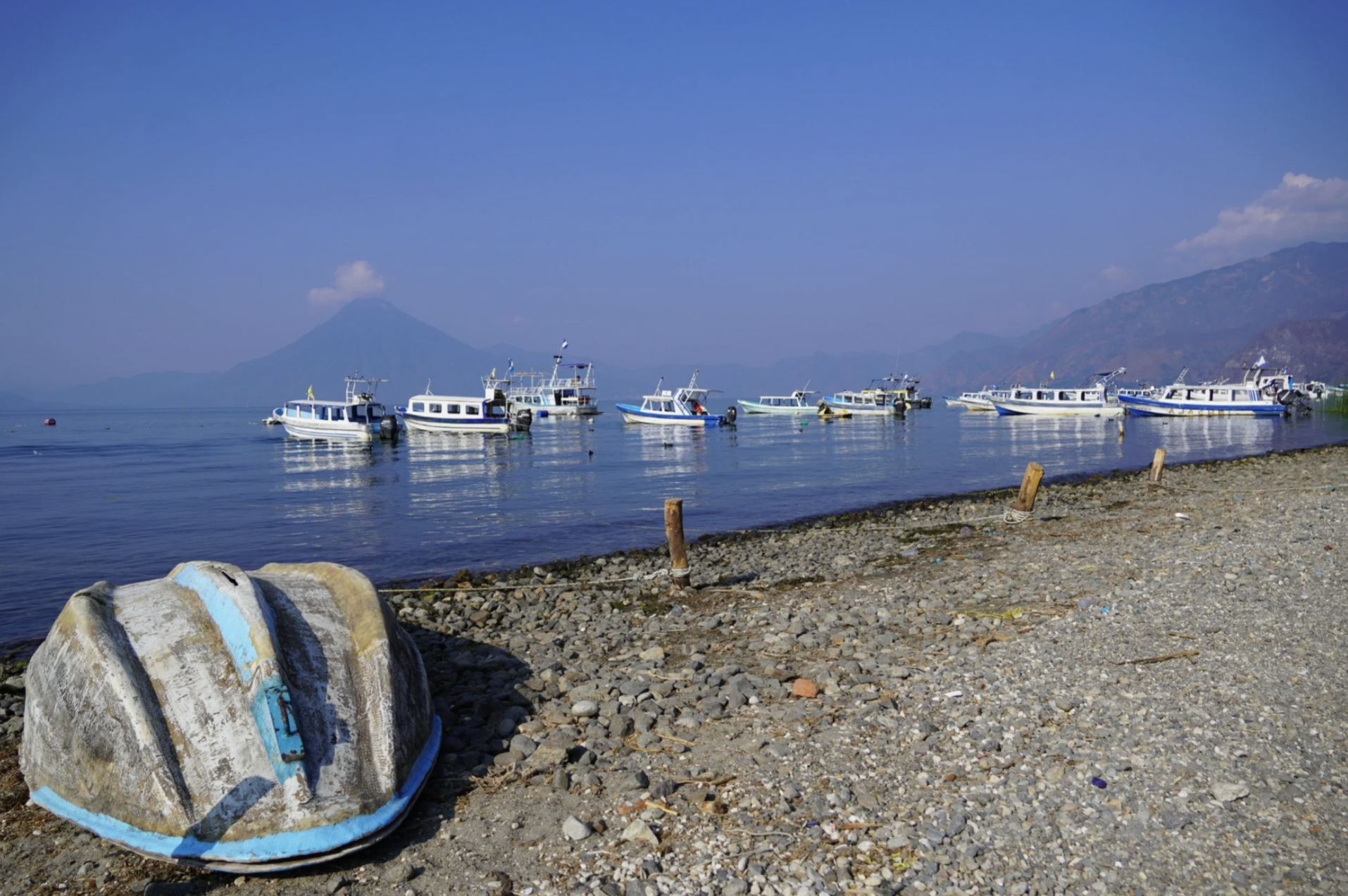 There are several boats sitting near the shore of Lake Atitlan in Guatemala with a volcano in the background.