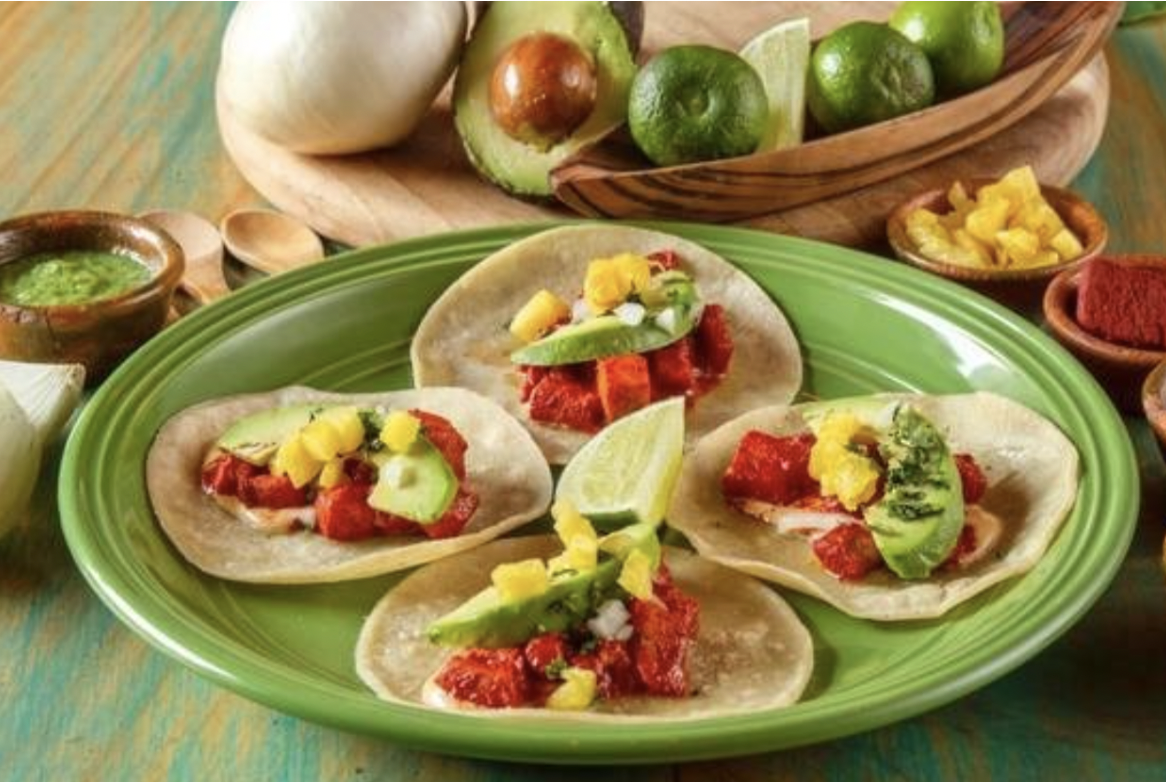 A plate of four tacos have veggies, avocado, and fresh salsa with a lime wedge in the middle surrounded by fresh local ingredients including pineapple, garlic, mojo, and an onion at one of the best restaurants in Antigua Guatemala.