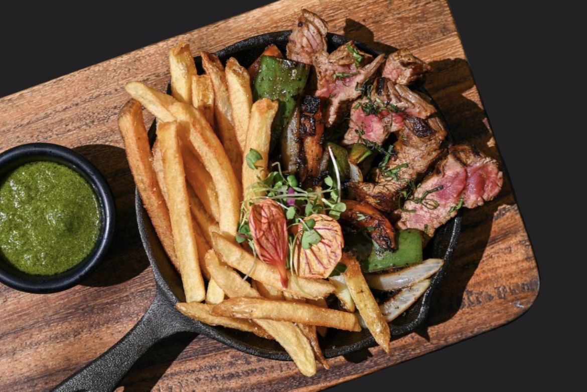 A medium rare steak and freshly cute french fries sit in a cast iron skillet with a side of sauce on a wooden board at Hector's Bistro, one of the the best restaurants in Antigua Guatemala
