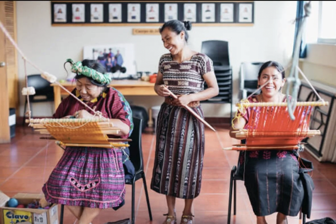 A woman stands over two Indigenous women as they are weaving on hand looms at Amigos de Santa Cruz, an organization you can donate to on Giving Tuesday