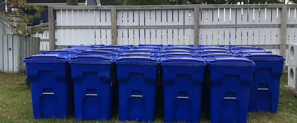 A long row of dark blue recycling bins with closed lids sitting on green grass in front of a white wooden fence. A long row of dark blue recycling bins with closed lids sitting on green grass in front of a white wooden fence.