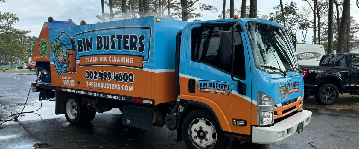 A blue and orange 'Bin Busters Trash Bin Cleaning' truck parked on wet asphalt, viewed from the side. A blue and orange 'Bin Busters Trash Bin Cleaning' truck parked on wet asphalt, viewed from the side.