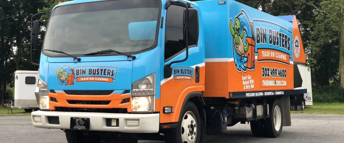 A Bin Busters trash bin cleaning truck with its turtle mascot, parked next to several green and black bins. A Bin Busters trash bin cleaning truck with its turtle mascot, parked next to several green and black bins.