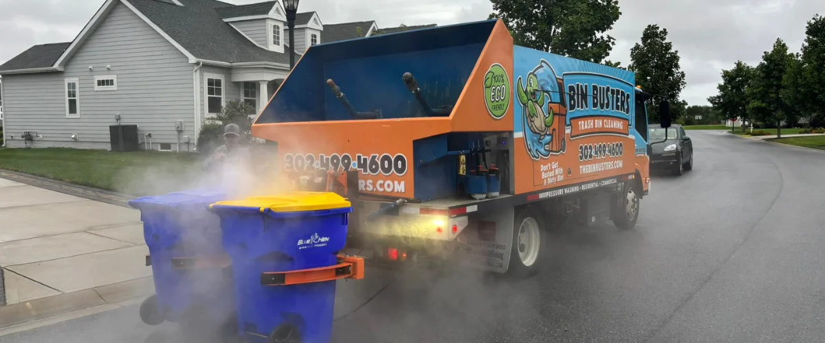 A Bin Busters trash bin cleaning truck cleaning two blue bins with steam on a residential street. A Bin Busters trash bin cleaning truck cleaning two blue bins with steam on a residential street.