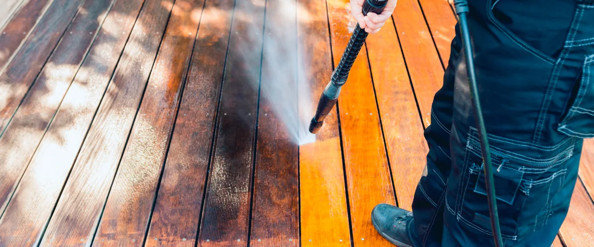 A person pressure washing a wooden deck, showing a clear distinction between the dirty and clean wood. A person pressure washing a wooden deck, showing a clear distinction between the dirty and clean wood.
