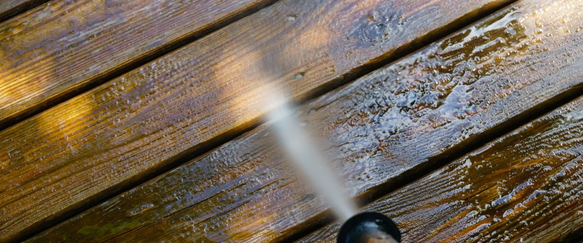 A close-up view of a wooden deck being power-washed, with a stream of water cleaning the planks. A close-up view of a wooden deck being power-washed, with a stream of water cleaning the planks.
