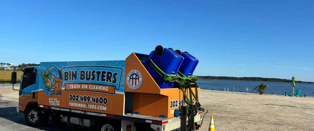 A Bin Busters trash bin cleaning truck with blue bins on its rear, parked on a sandy waterfront with water and trees in the background. A Bin Busters trash bin cleaning truck with blue bins on its rear, parked on a sandy waterfront with water and trees in the background.