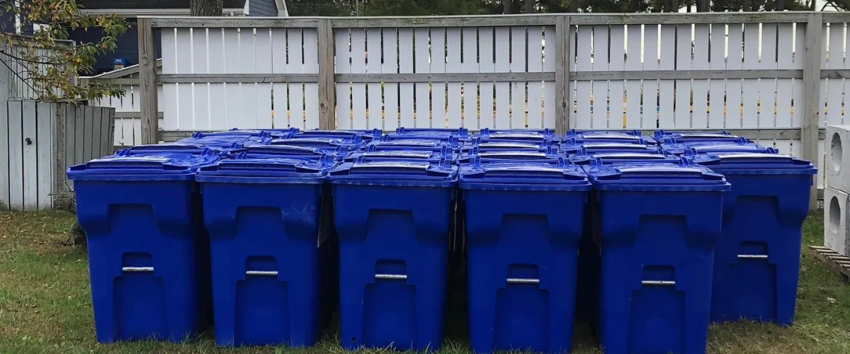A long row of blue recycling bins with lids stands on green grass in front of a white wooden fence. A long row of blue recycling bins with lids stands on green grass in front of a white wooden fence.