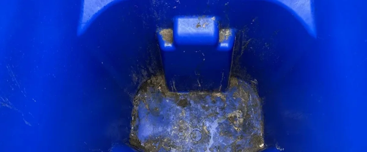 A close-up, top-down view inside a dirty blue recycling bin with accumulated grime and residue at the bottom. A close-up, top-down view inside a dirty blue recycling bin with accumulated grime and residue at the bottom.