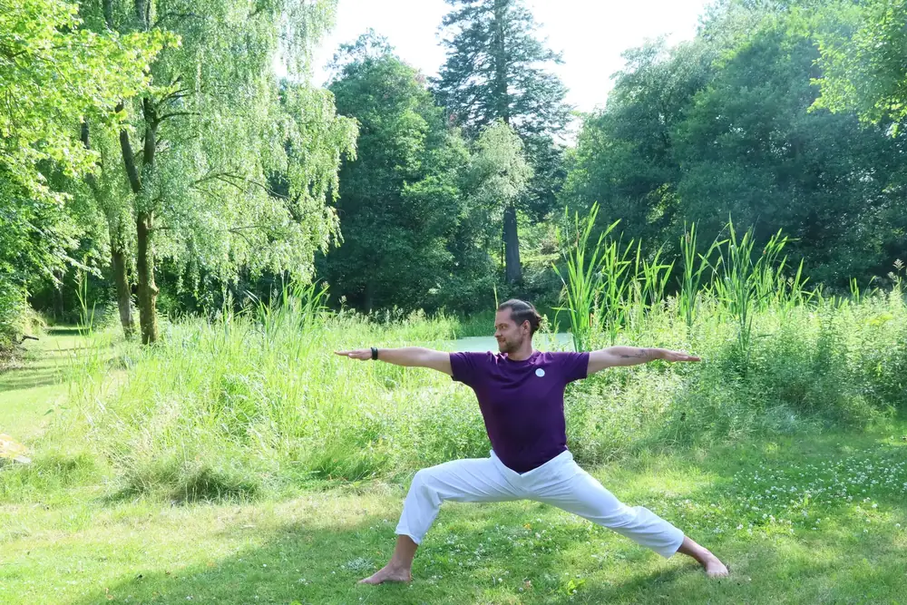 Aurimas Savickas in warrior pose (Virabhadrasana II) on a green meadow by a pond