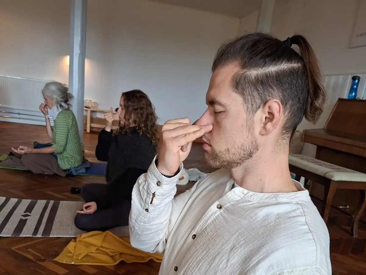 Aurimas Savickas doing alternate-nostril breathing (nadi shodhana) in a yoga hall, with two women practising behind him