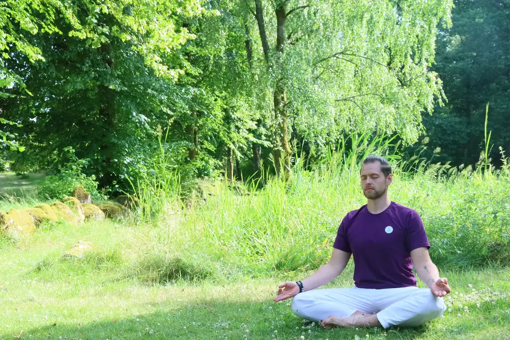 Aurimas Savickas meditating cross-legged with closed eyes in a meadow in front of a birch tree
