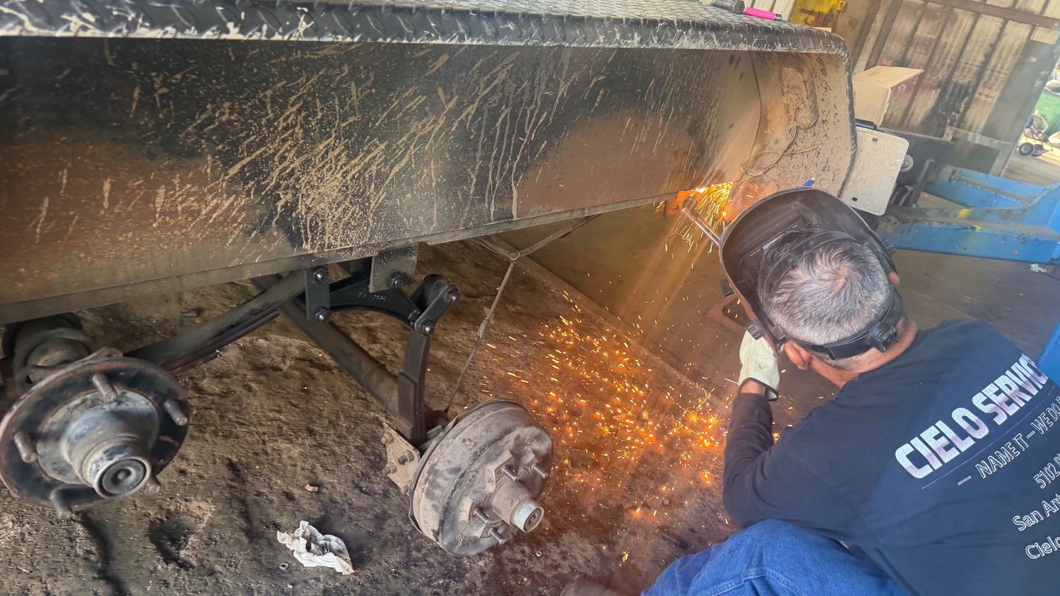 Technician welding a trailer frame repair under a dump trailer at Mr. Cielo Trailers in San Antonio
