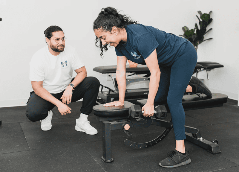 A patient performing dumbbell press on a bench & the trainer looking at her in Vancouver, Surrey & Langley