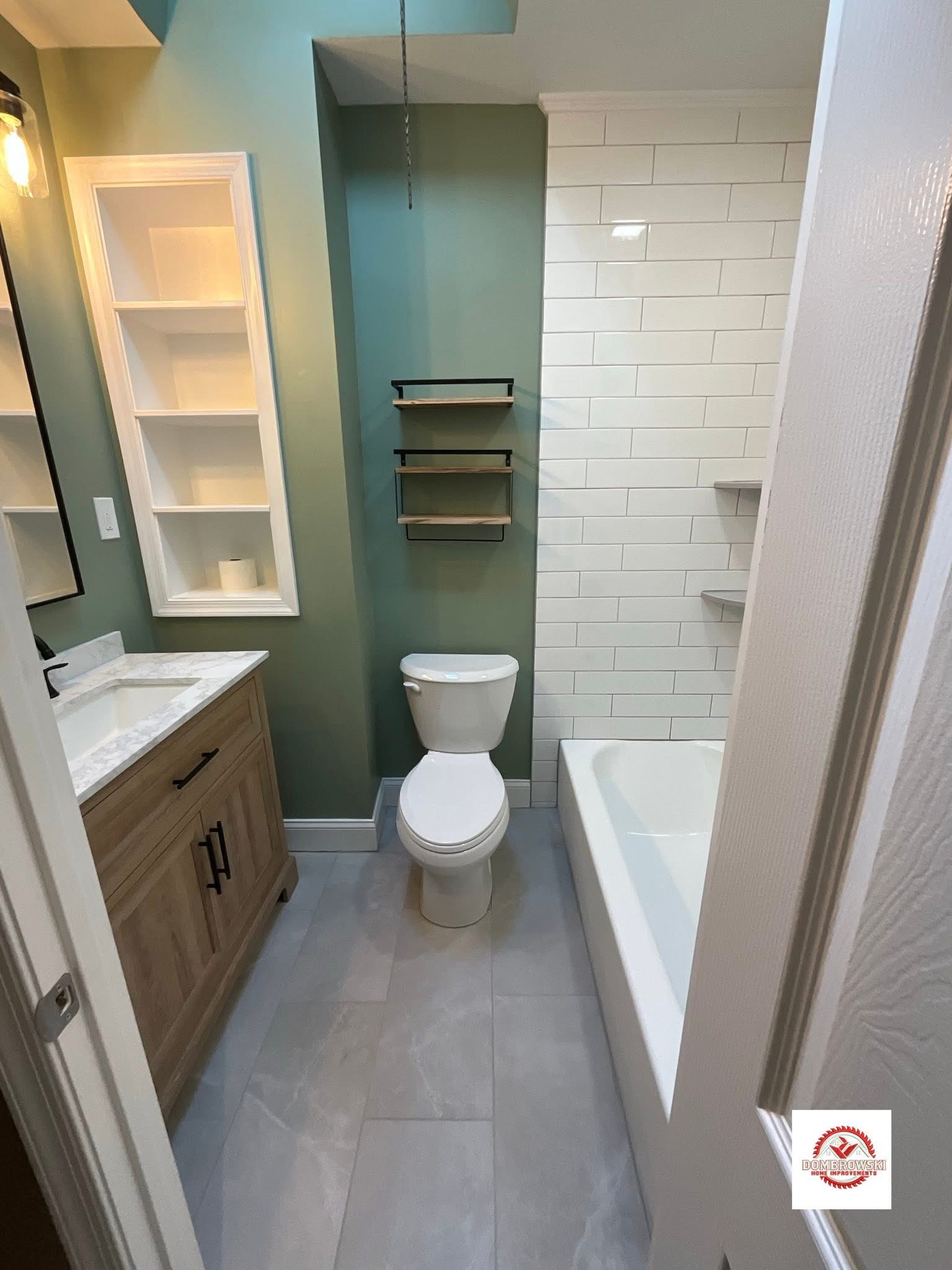 Sage green bathroom full view: tub surround, natural wood vanity and built-in shelving