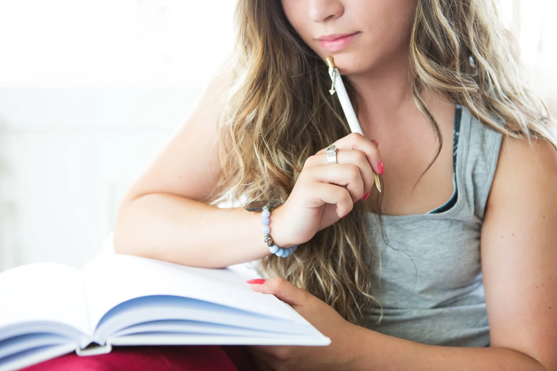 A young woman holding a pen to her chin as she pauses thoughtfully over an open notebook at a table