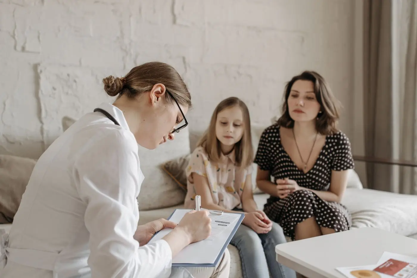 A counselor taking notes while a mother and her young daughter talk with her on a sofa in a bright office