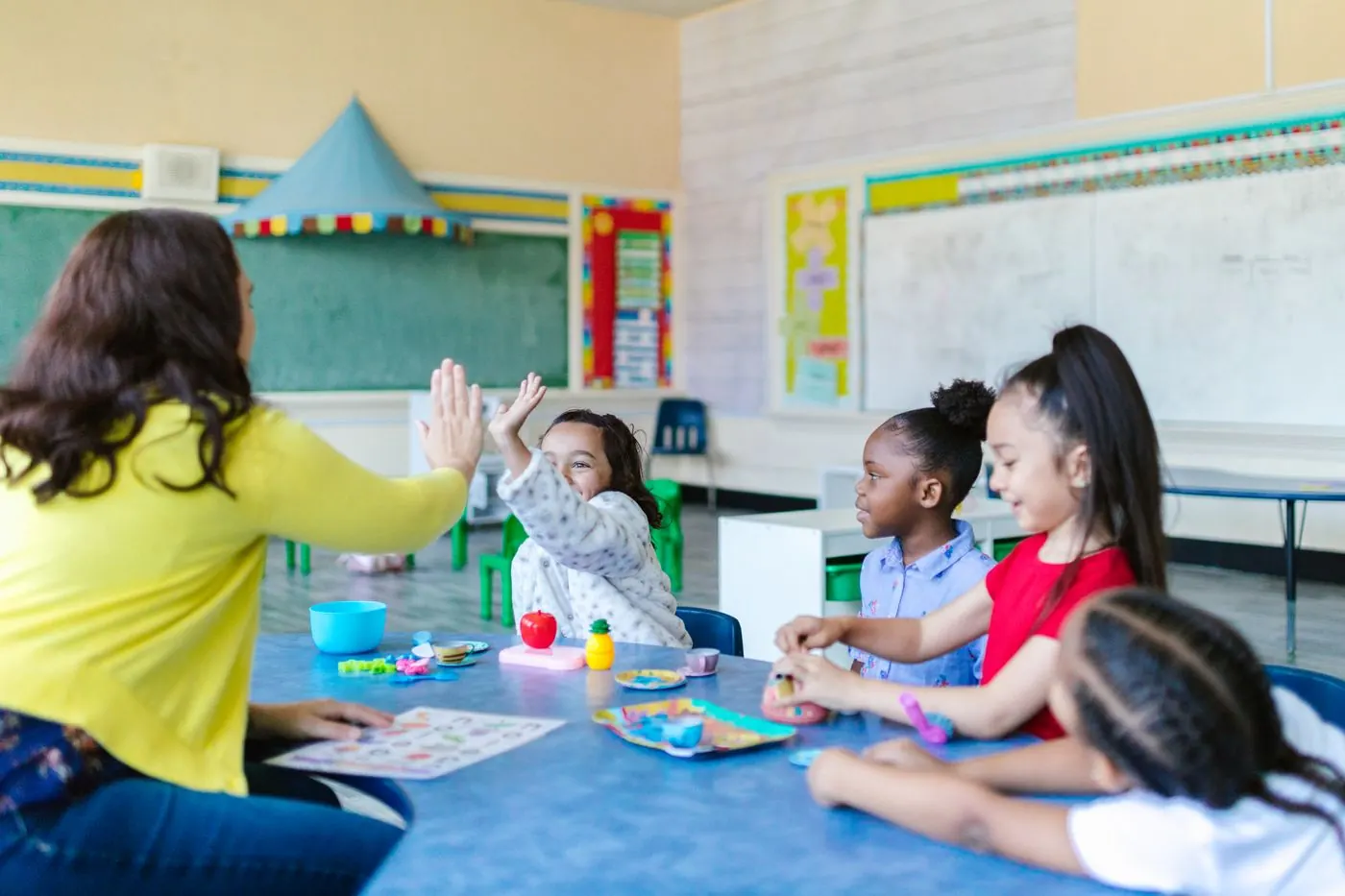 A smiling teacher leading a diverse group of happy kids around a classroom table, one girl raising her hand