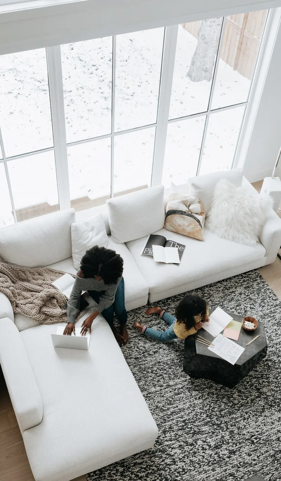 A mother on a laptop on the sofa while her daughter draws at the coffee table.