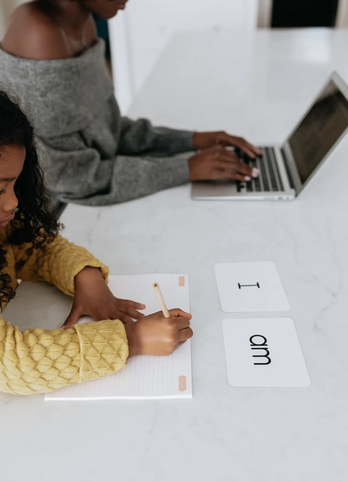 A child writing in her Power Journal beside word cards reading 'I am' while her mother works on a laptop.