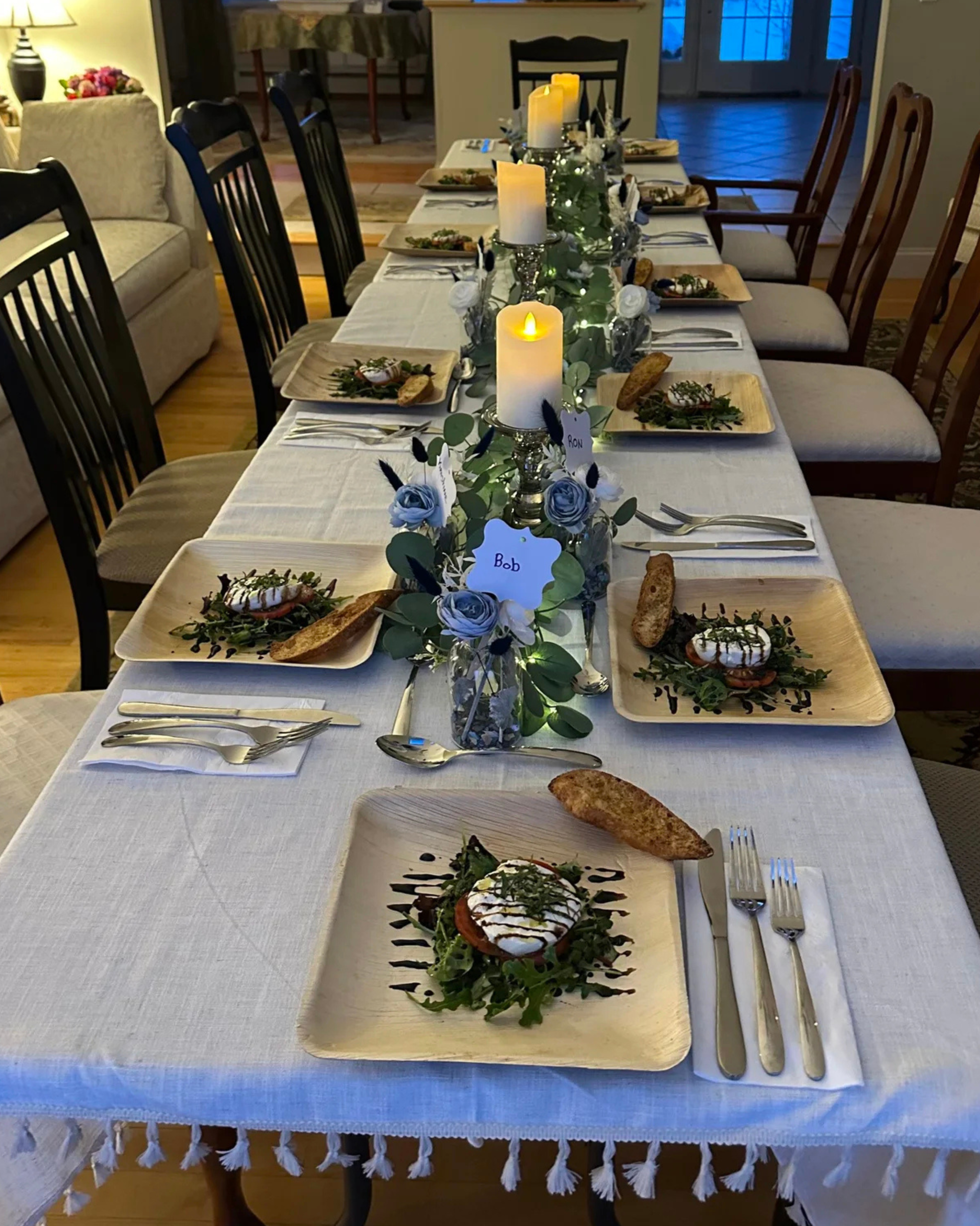 A long, beautifully set dinner table featuring elegant greenery, lit candles, and individually plated meals for an intimate home celebration or micro-wedding.