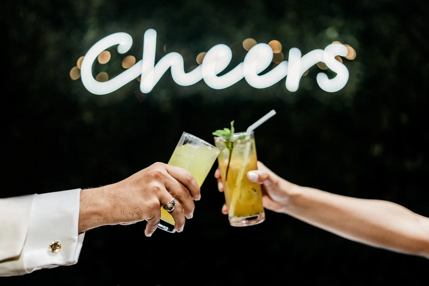 Los novios brindan con dos c&oacute;cteles frente a un letrero de ne&oacute;n que dice Cheers