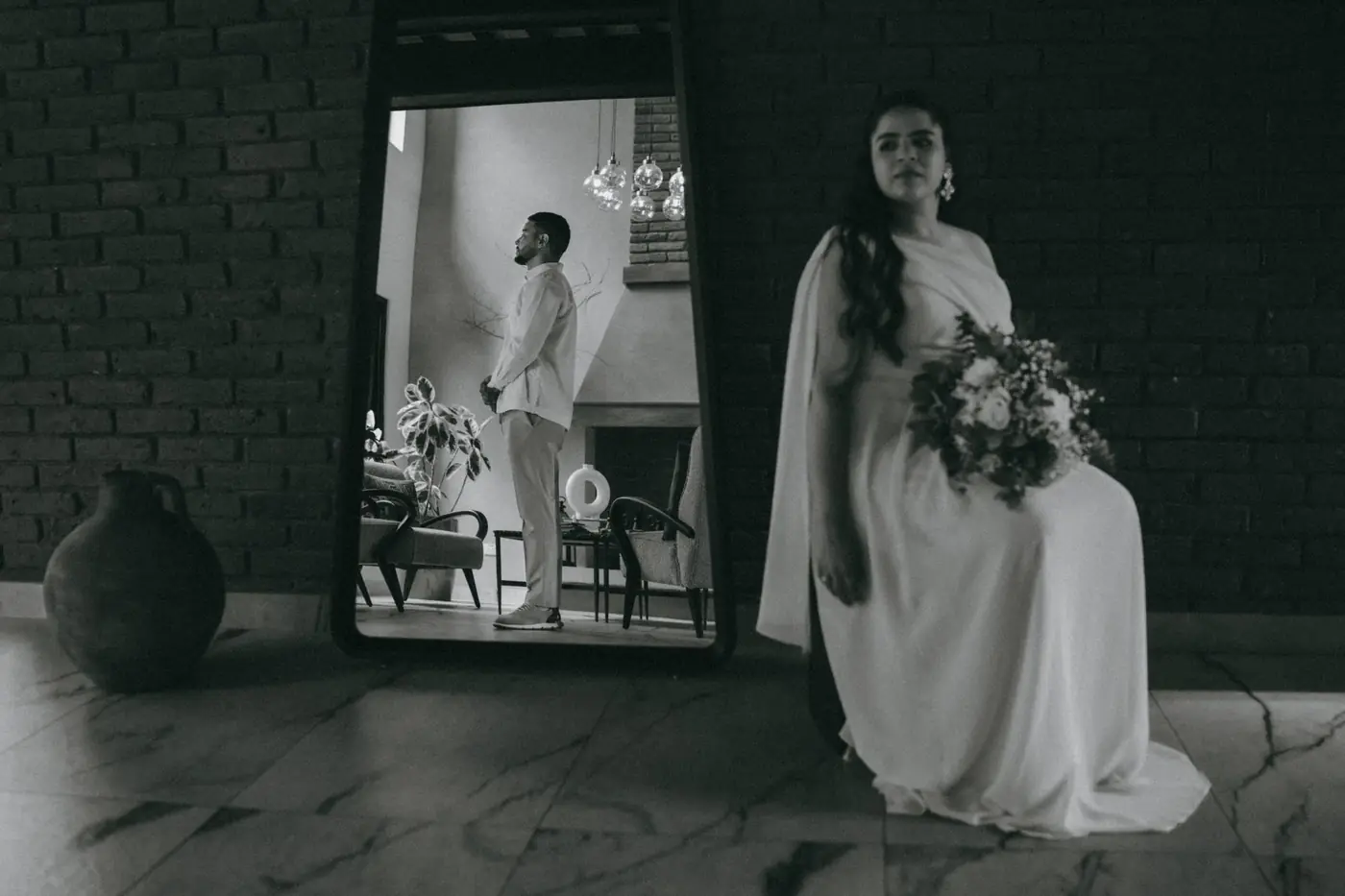 La novia sentada junto a un espejo en el que se refleja el novio de pie, esperando