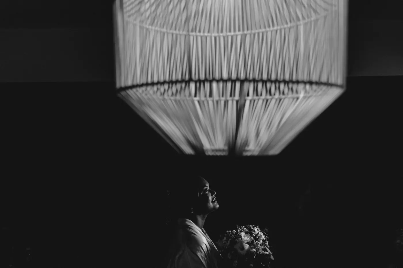 La novia mira hacia arriba bajo una gran l&aacute;mpara tejida, con el ramo en las manos
