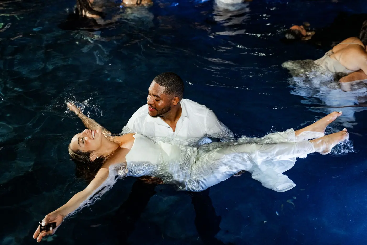 La novia flota en la piscina de noche, sostenida por el novio, riendo con los brazos abiertos