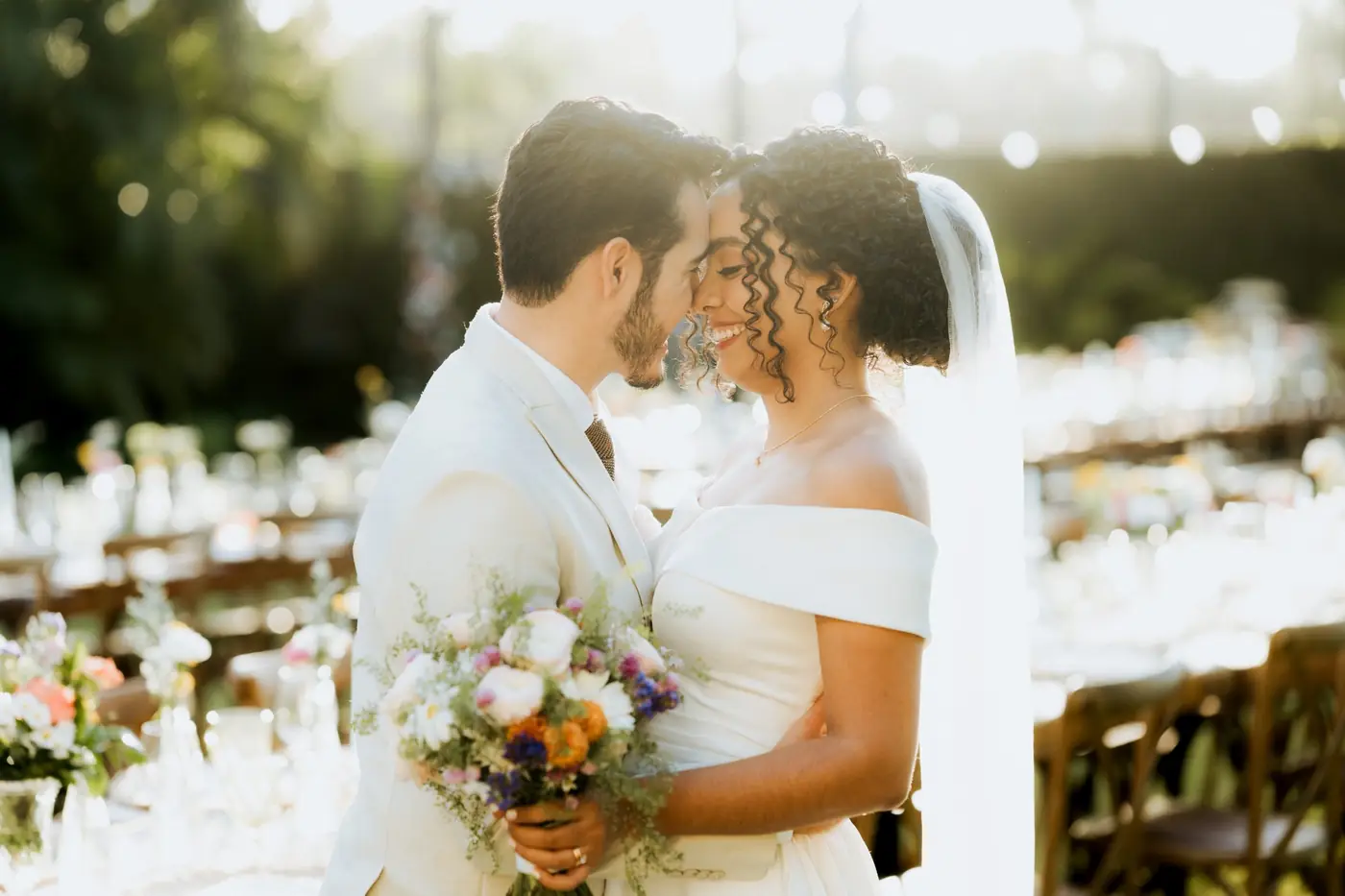 Los novios frente con frente, sonriendo a contraluz entre las mesas del banquete