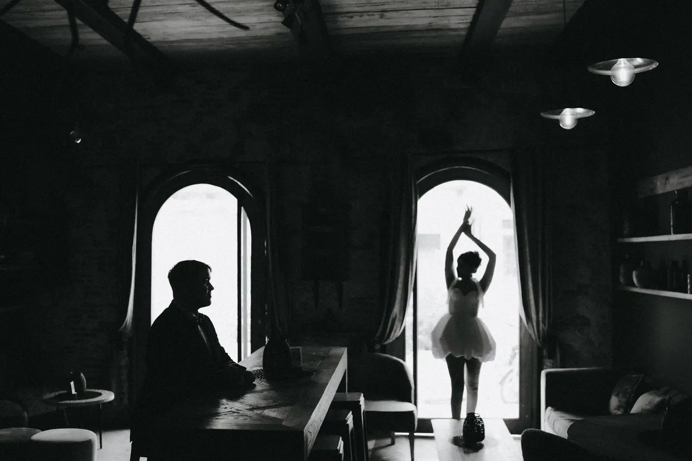 Silueta de la novia en pose de ballet frente a una ventana en arco, con el novio sentado a la izquierda