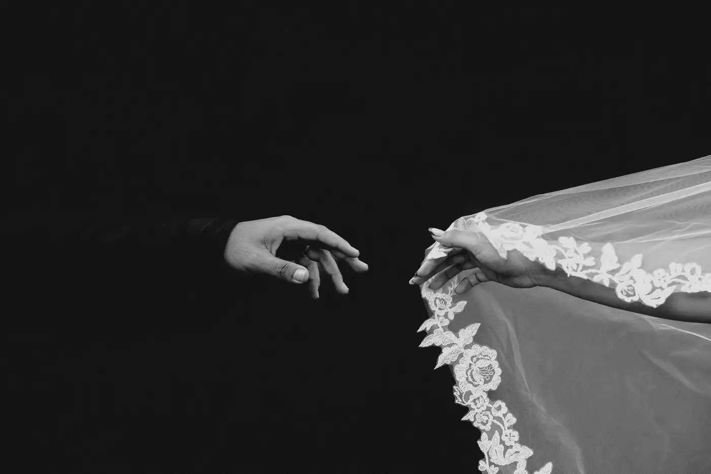 La mano del novio y la de la novia, cubierta por el velo, casi se tocan sobre fondo negro