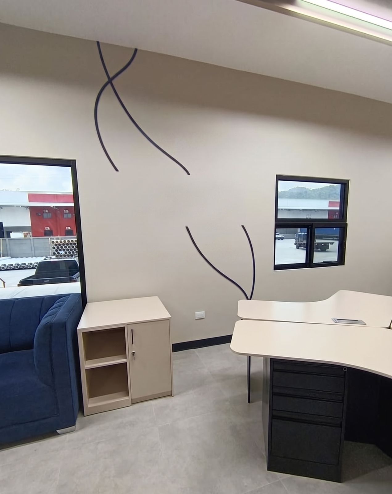 Oficina de gerencia con sala de estar, muro beige con grafismo curvo y ventana al patio industrial