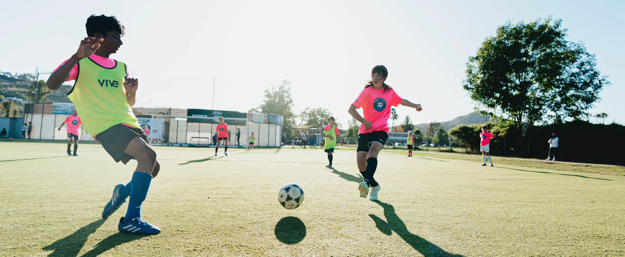 Two young players in a Soro Futbol training session at golden hour, one in a pink Soro jersey and one in a lime bib, both running at a ball on the turf as a group session carries on behind them.