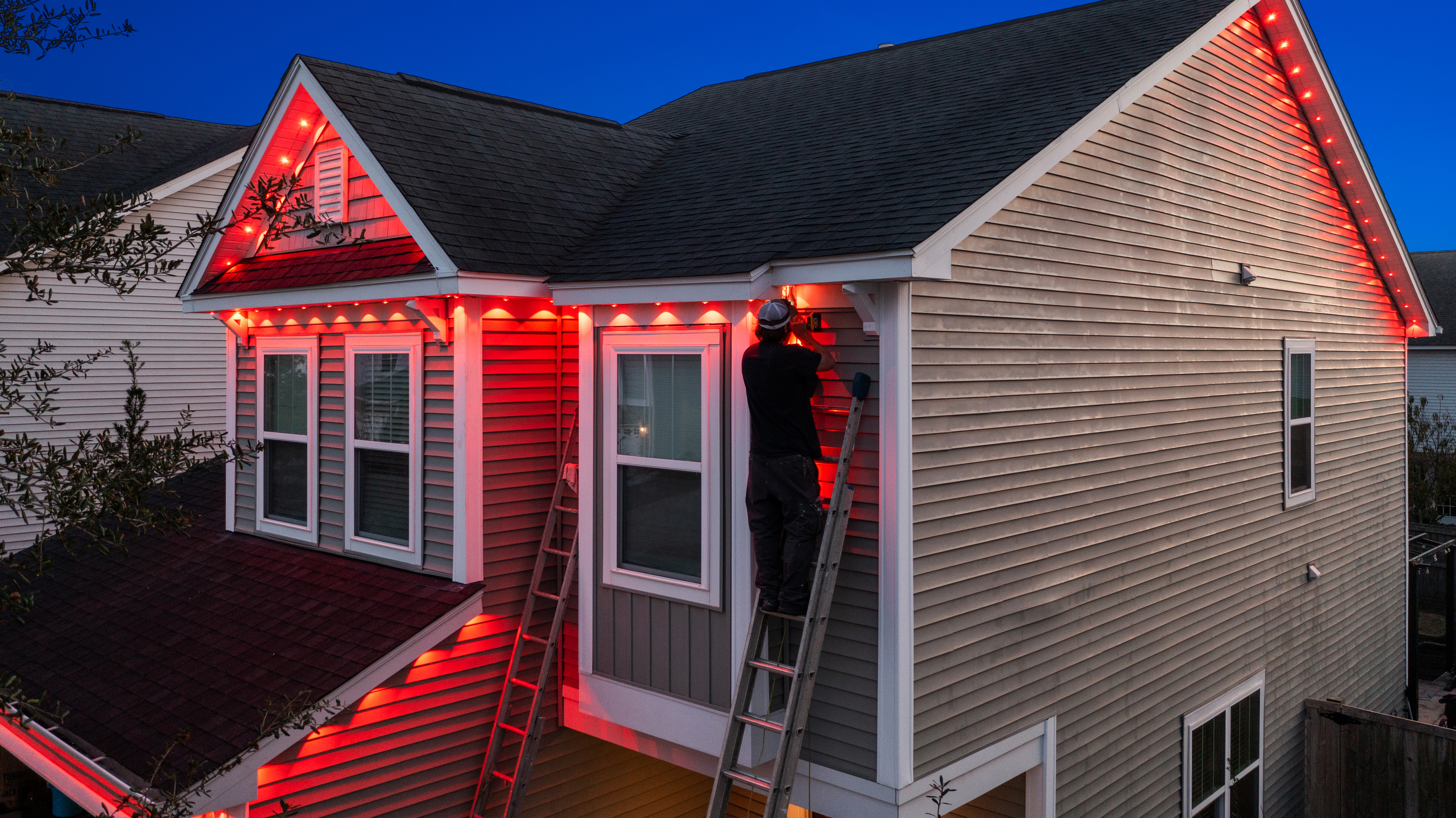 Professional Technician installing red permanent outdoor LED lights along the roofline of a two-story home at dusk.