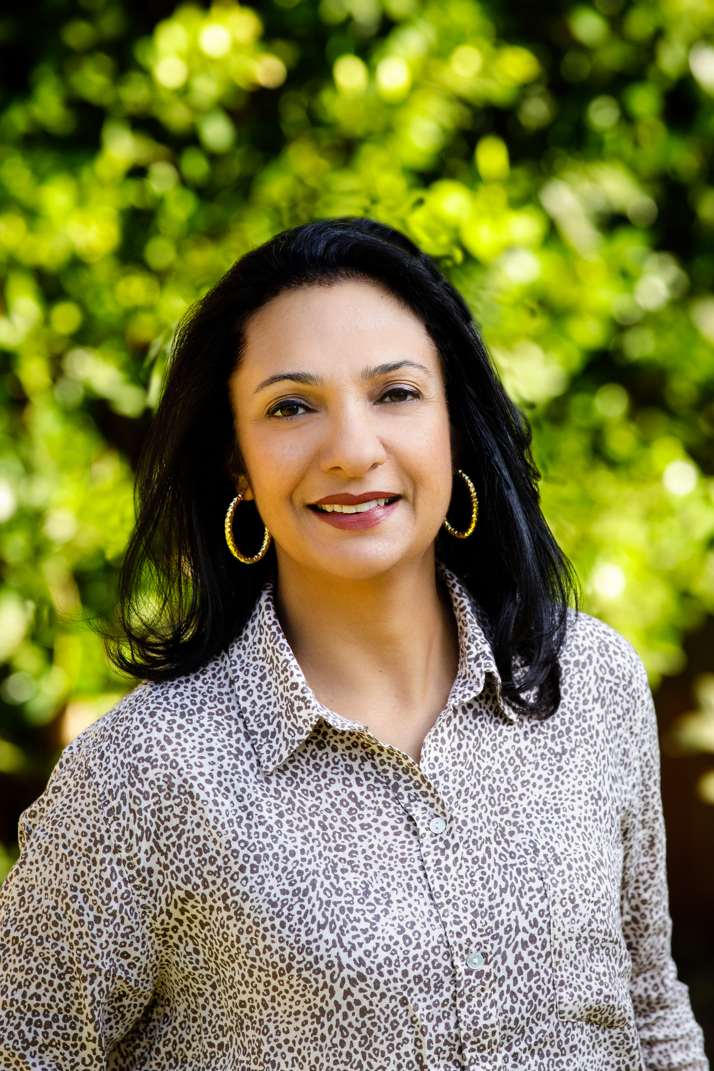 Professional outdoor headshot of a woman wearing a patterned blouse, standing in front of softly blurred green foliage.