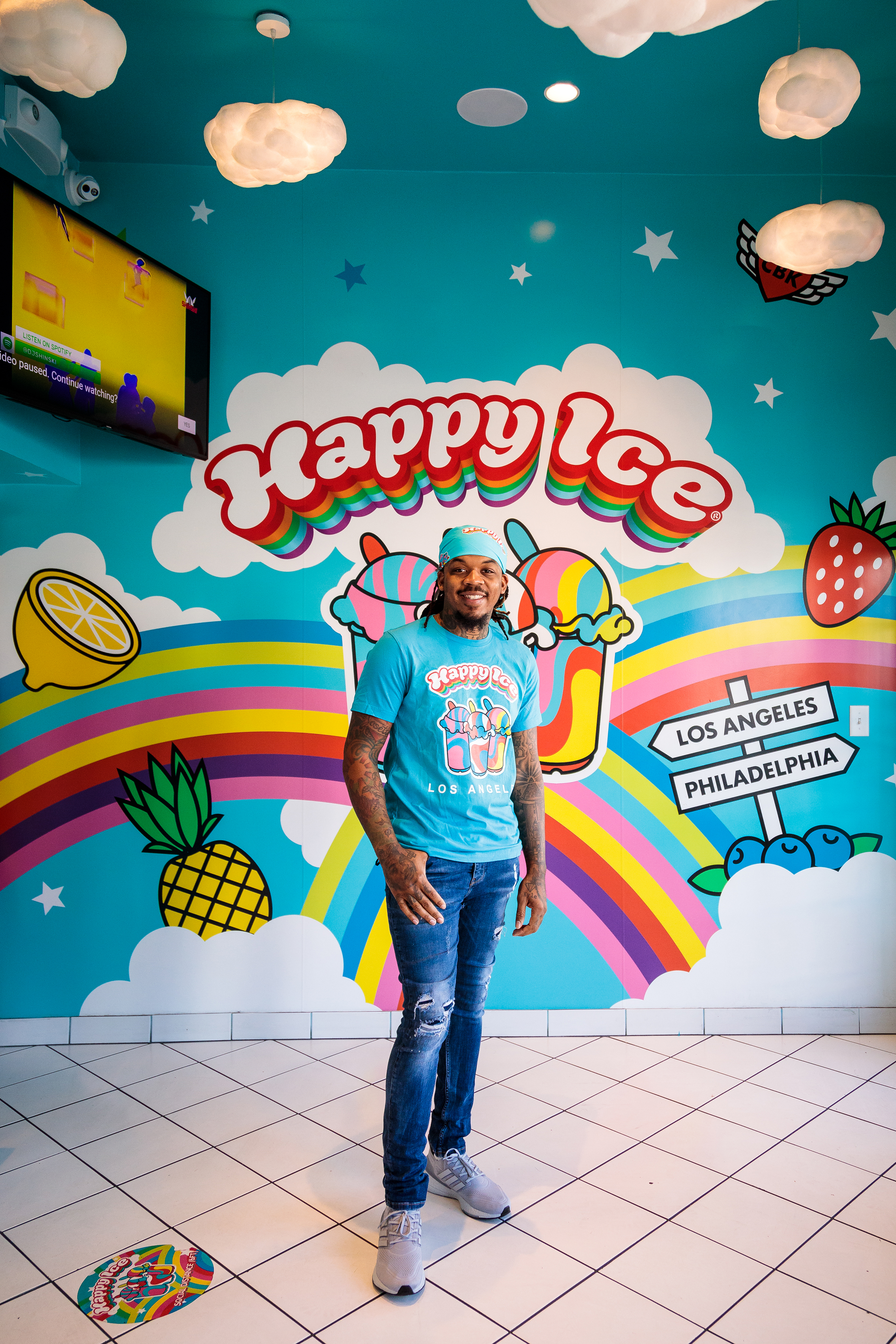 Man at Happy Ice storefront. CEO Lemeir Mitchell at Happy Ice wearing branded Happy Ice t-shirt standing in front of a colorful dessert shop mural with rainbows, fruit, and playful signage. Branding portrait in a vibrant retail environment.