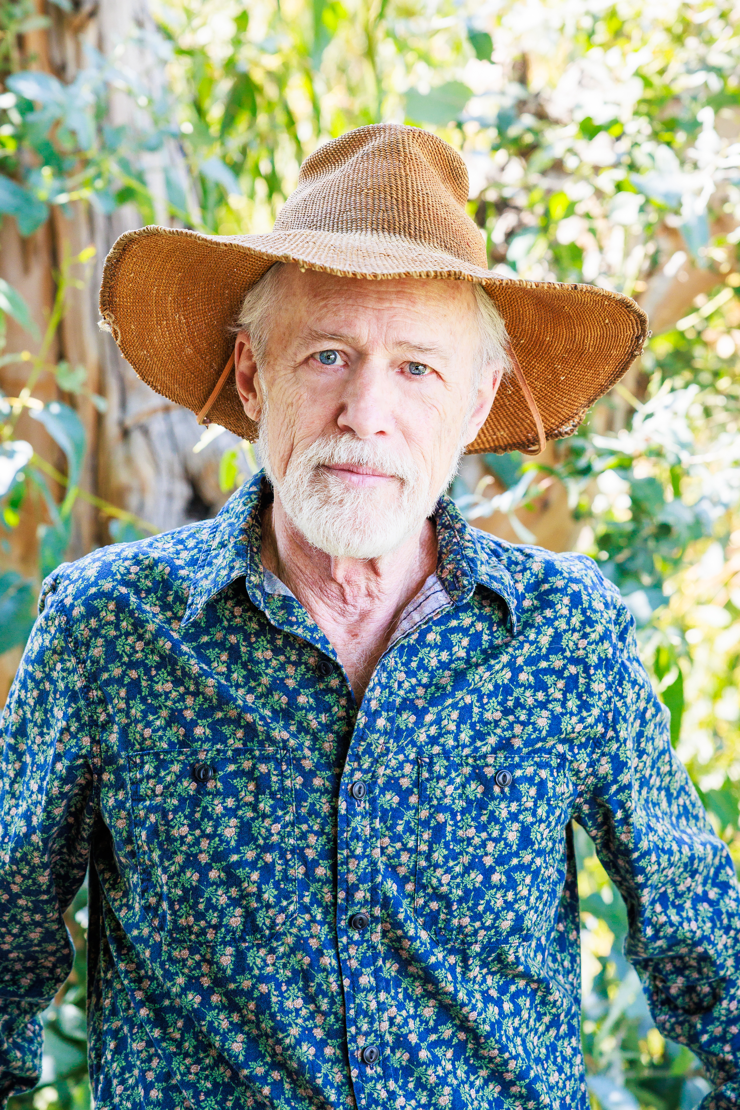 James Goodwin wearing a straw hat and patterned shirt standing outdoors in soft natural light, surrounded by greenery.