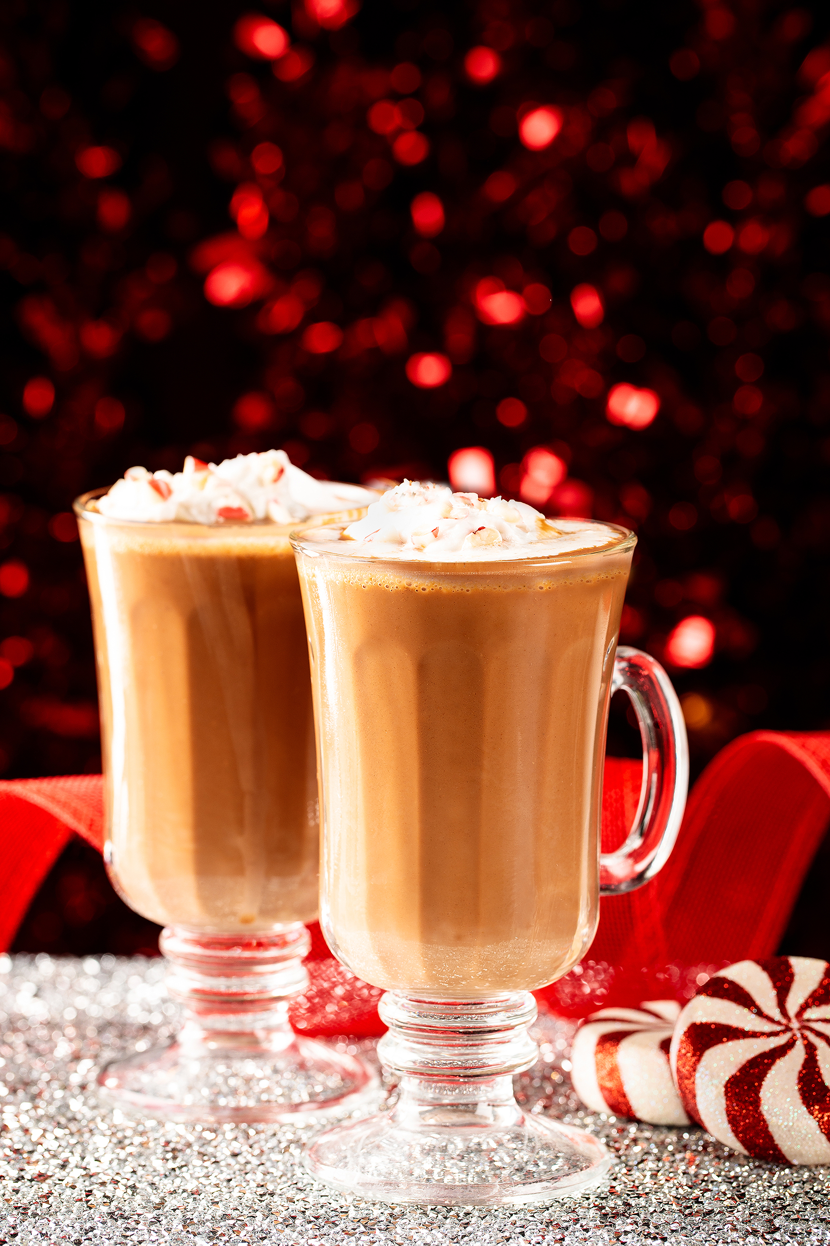 Two glasses of vegan peppermint hot cocoa topped with whipped cream, photographed with glittering red holiday décor.