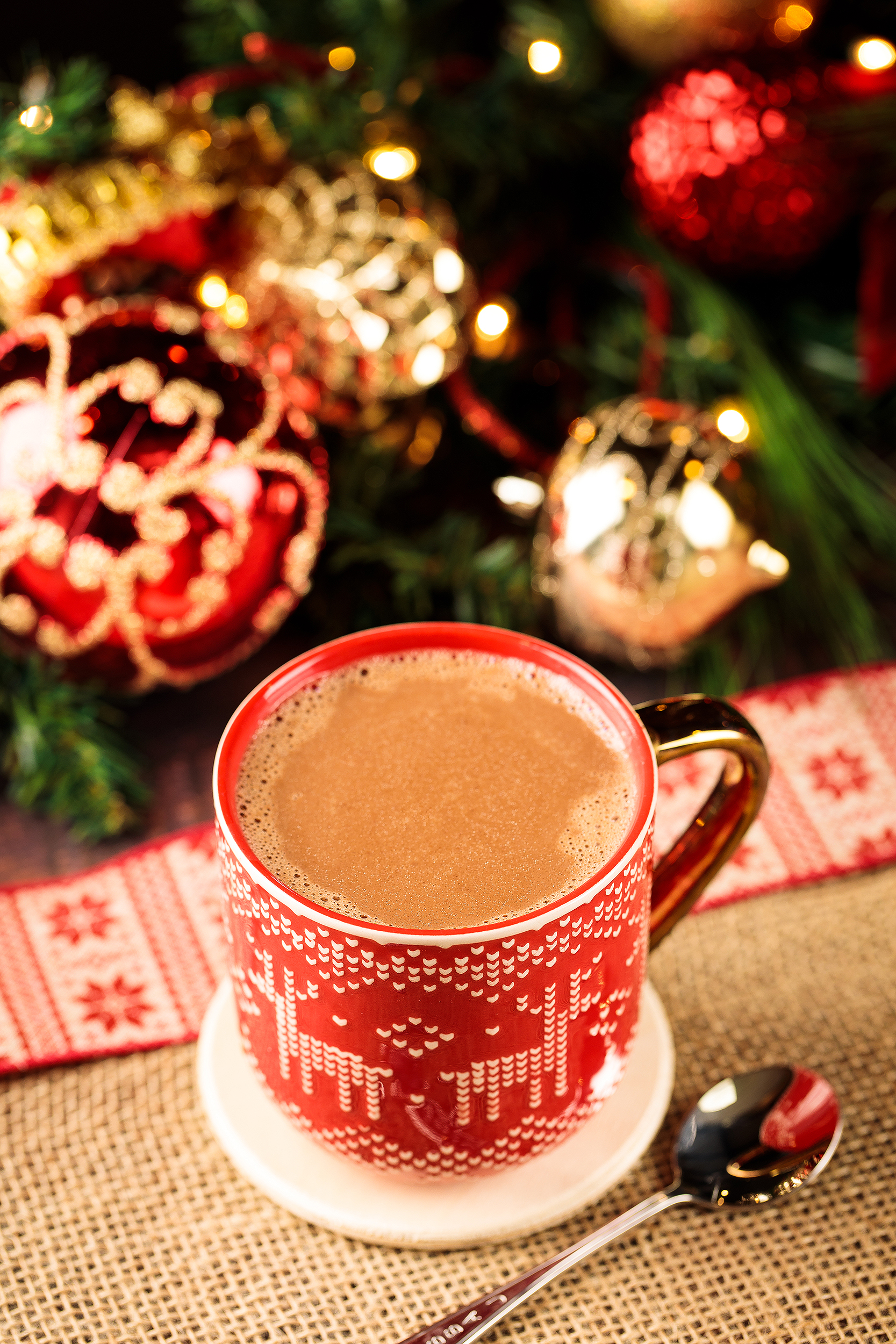 Vegan hot cocoa in a red knit-pattern mug surrounded by Christmas ornaments, styled for a holiday cookbook photoshoot.