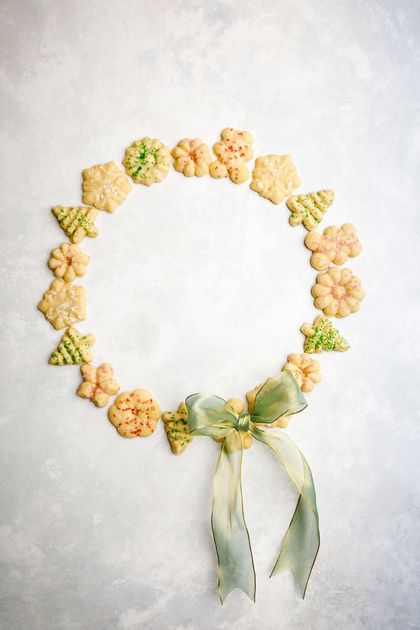 Assorted vegan spritz cookies arranged in a Christmas wreath shape with a gold ribbon, photographed for a holiday cookbook.