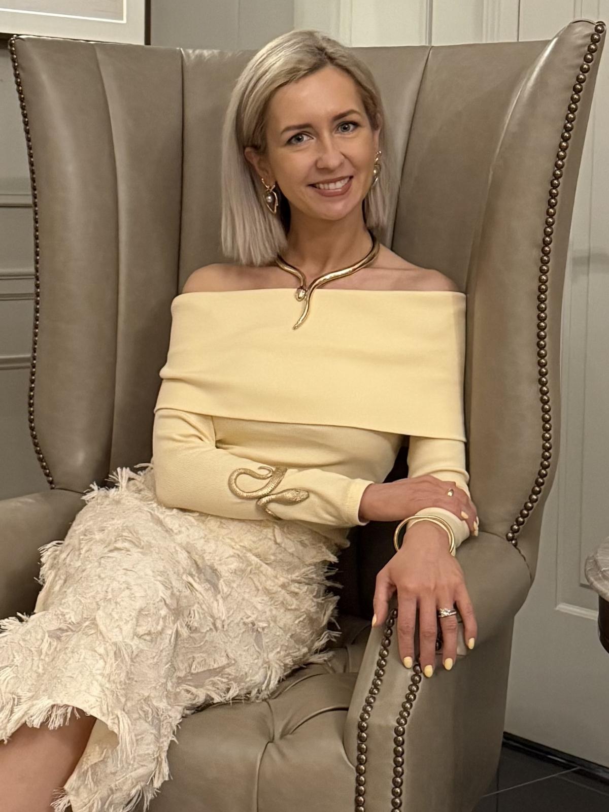 Lena Aspette seated in an upholstered wing-back chair, wearing a cream off-shoulder top with a feather-textured skirt and gold snake necklace