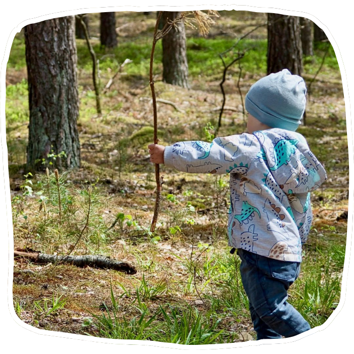 A young child in a blue beanie and patterned shirt walks through the woods holding a long, thin branch.
