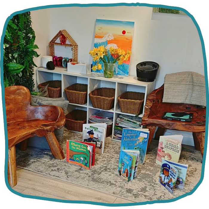 A children’s reading nook has a low white cubby shelf with baskets and books, two chairs, a flower painting, and books displayed on the rug. at nursery in Castlemead