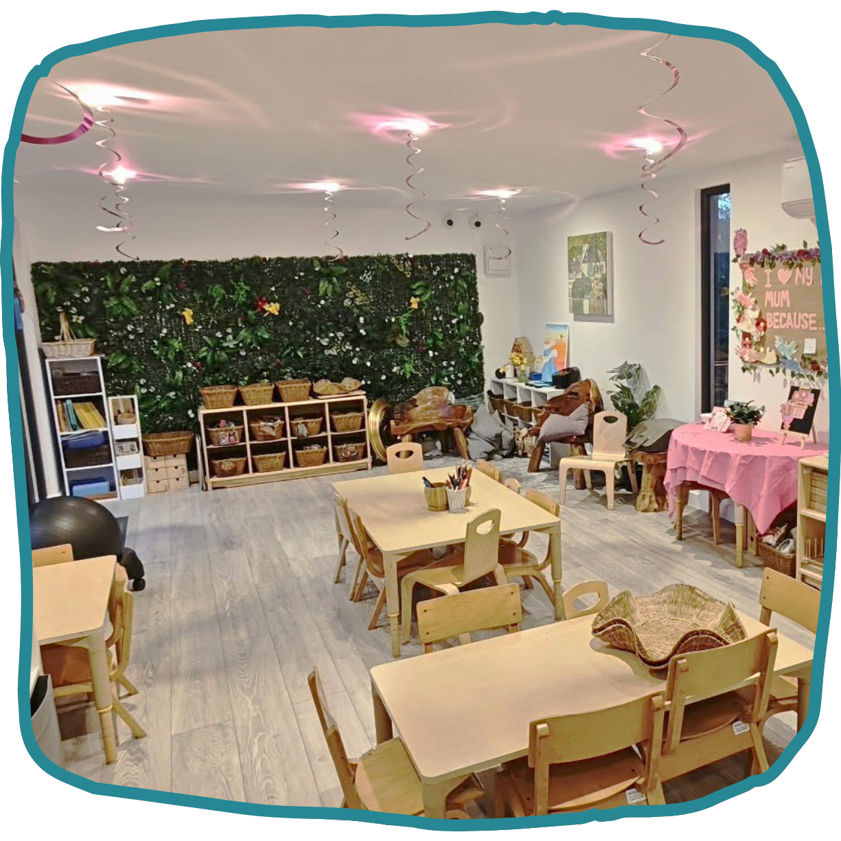 A children’s room with low wooden tables and chairs, baskets on shelves against a leafy wall, ceiling streamers in nursery at Morden