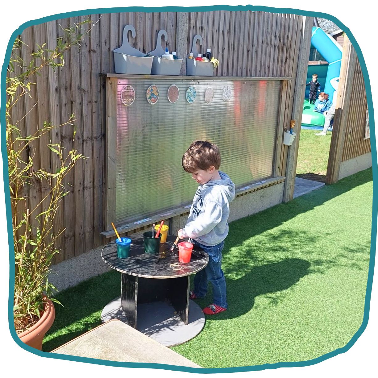 A boy stands at a round outdoor table with colorful cups and paintbrushes beside a wooden water-play wall at nursery in Morden