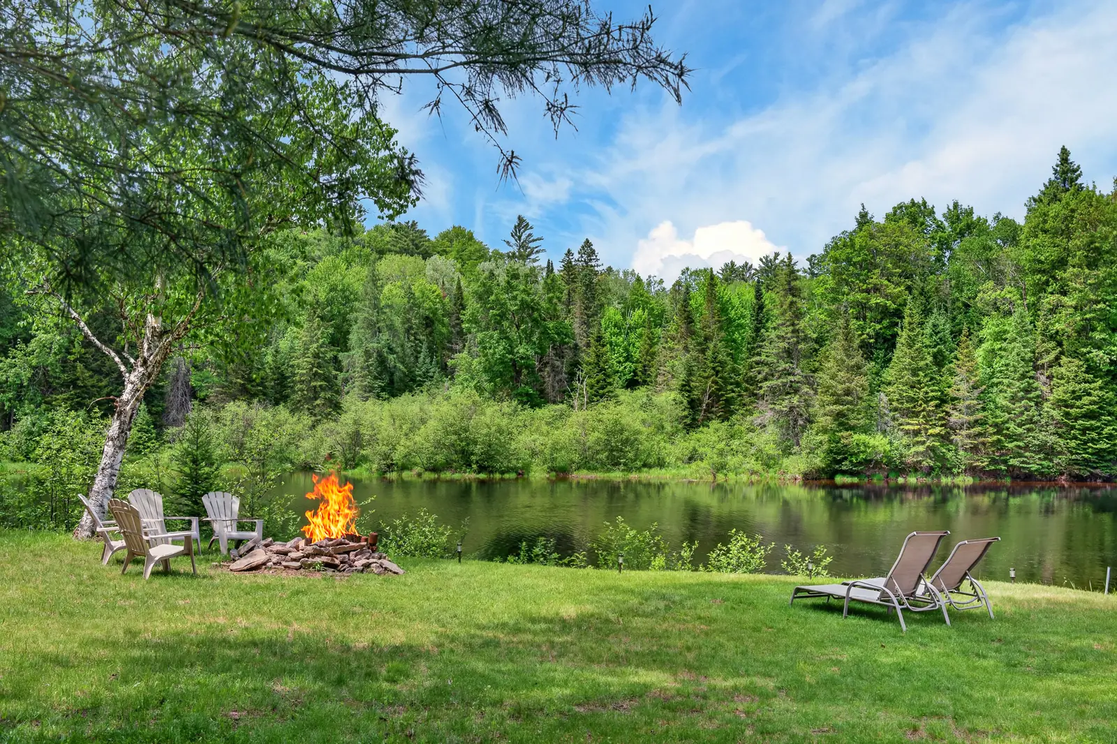 Lit fire pit and loungers on The Mara riverside lawn, with forest across the water