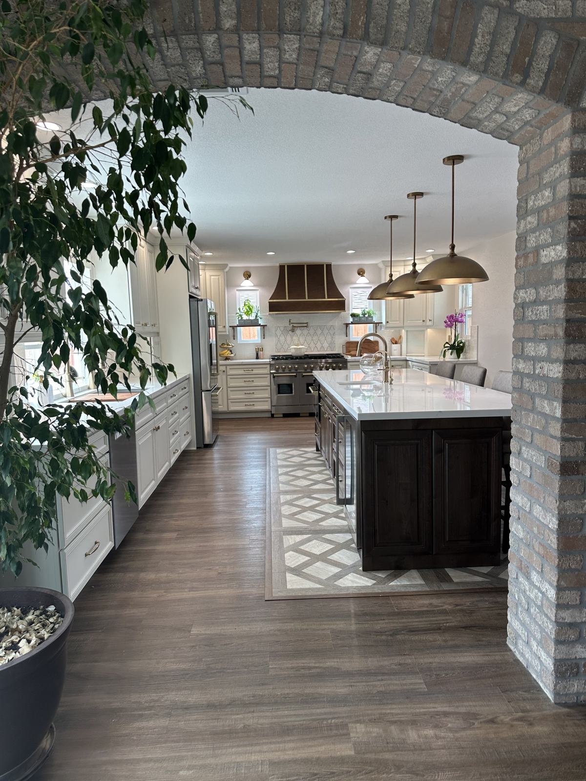Remodeled kitchen seen through a brick archway with white cabinets, dark wood island, quartz counters and brass pendant lights