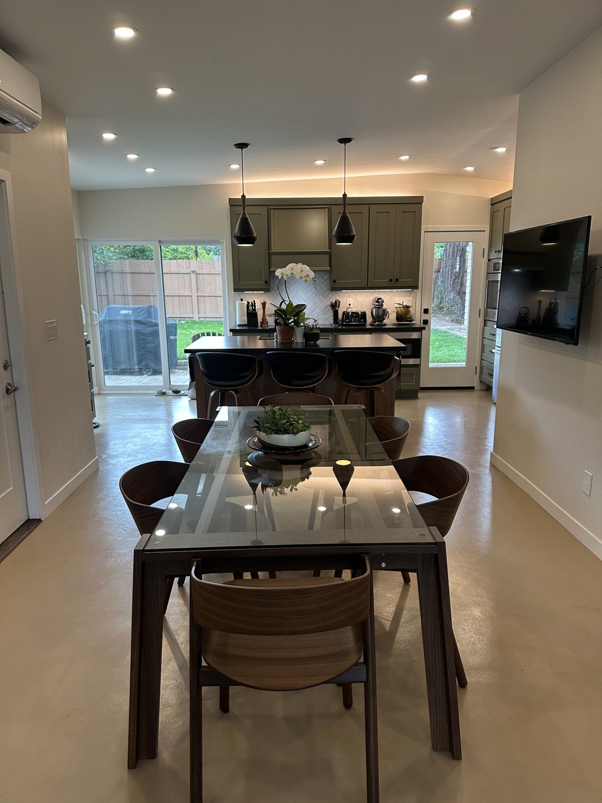 Remodeled kitchen viewed from the dining table with sage green cabinets, black pendants and glass dining table